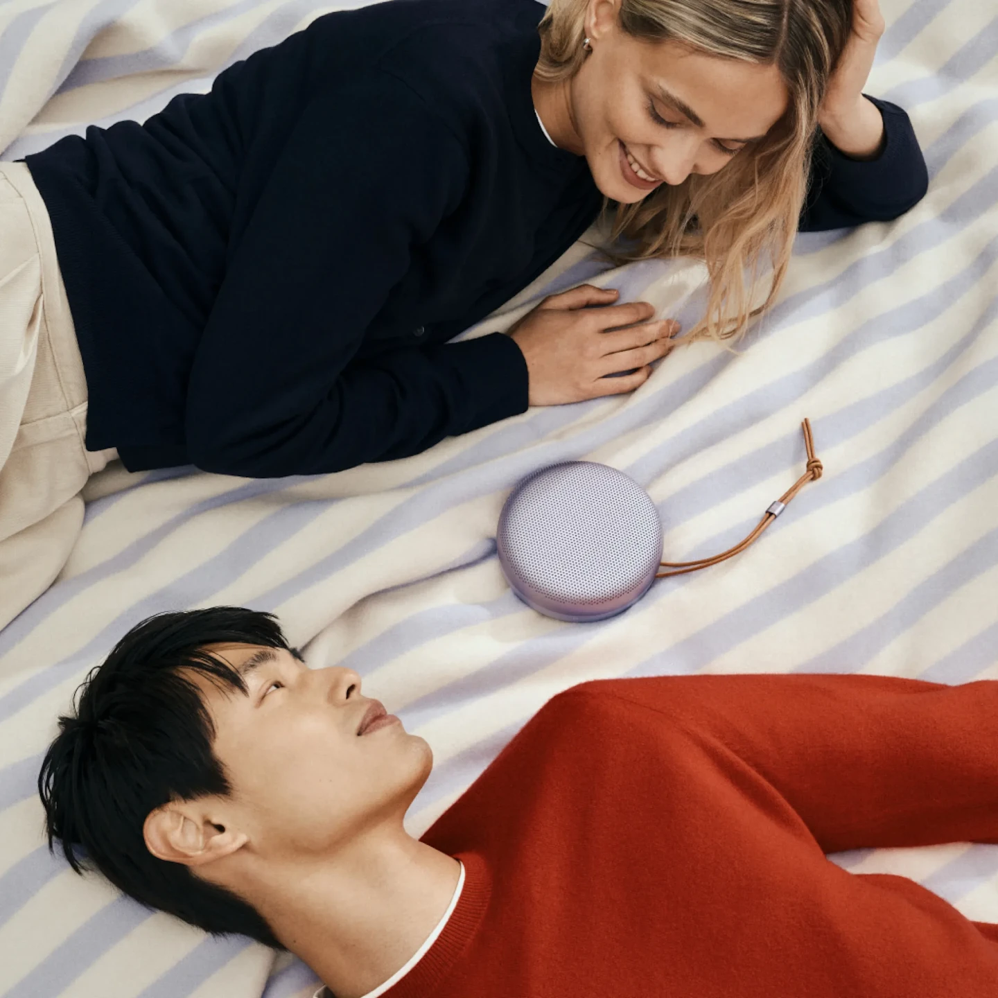 A couple lying face-to-face on a picnic mat with a Bang & Olufsen Beosound A1 in Lavender placed between them, sharing a relaxed moment through music.