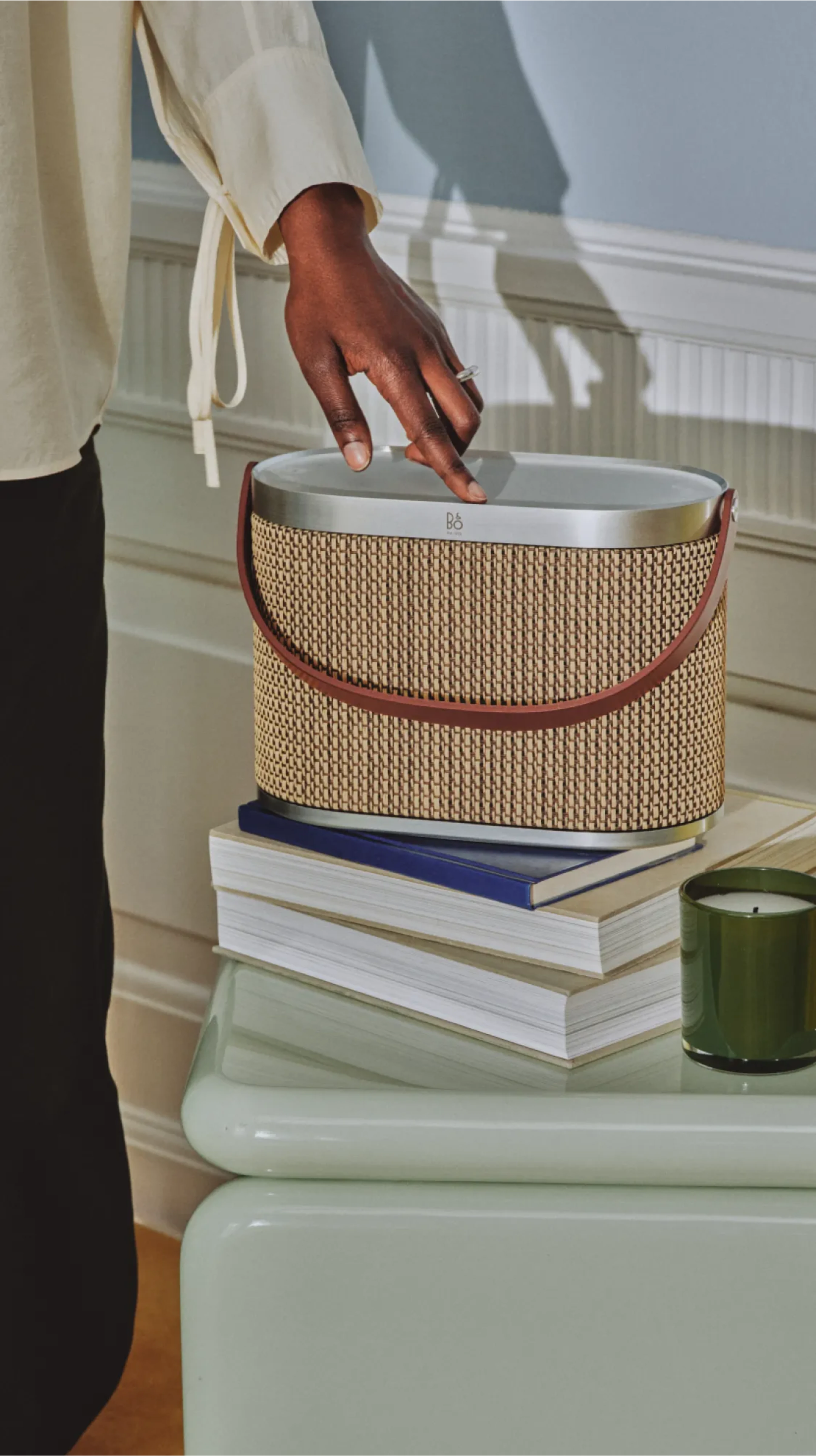 Woman using Beosound A5 in the variant Century Weave. The speaker is on a side table with books and candle.