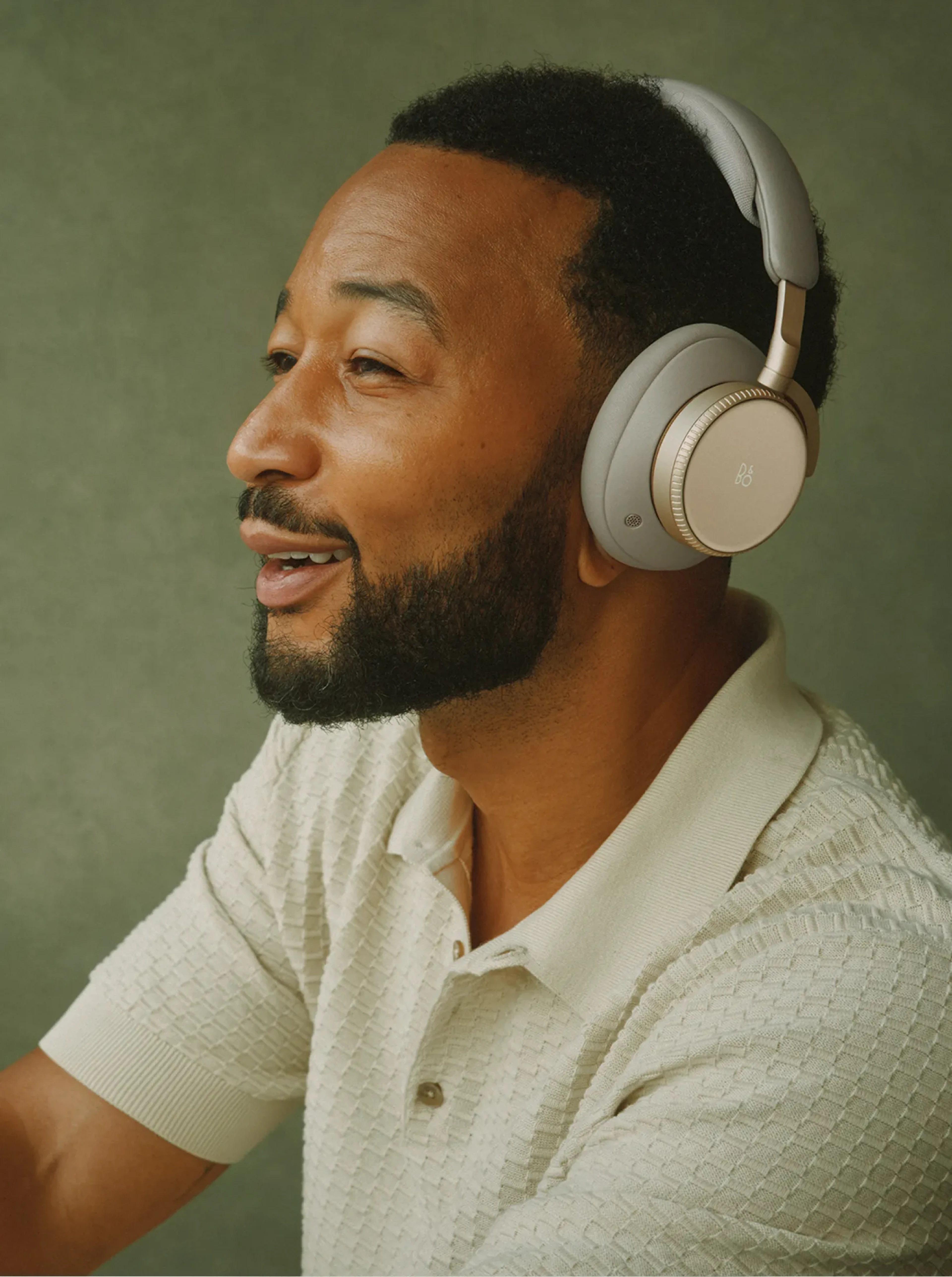 John Legend depicted wearing Bang & Olufsen Beoplay H100 over-ear headphones in the Hourglass Sand colorway, photographed against a green background in a relaxed, music-listening pose.