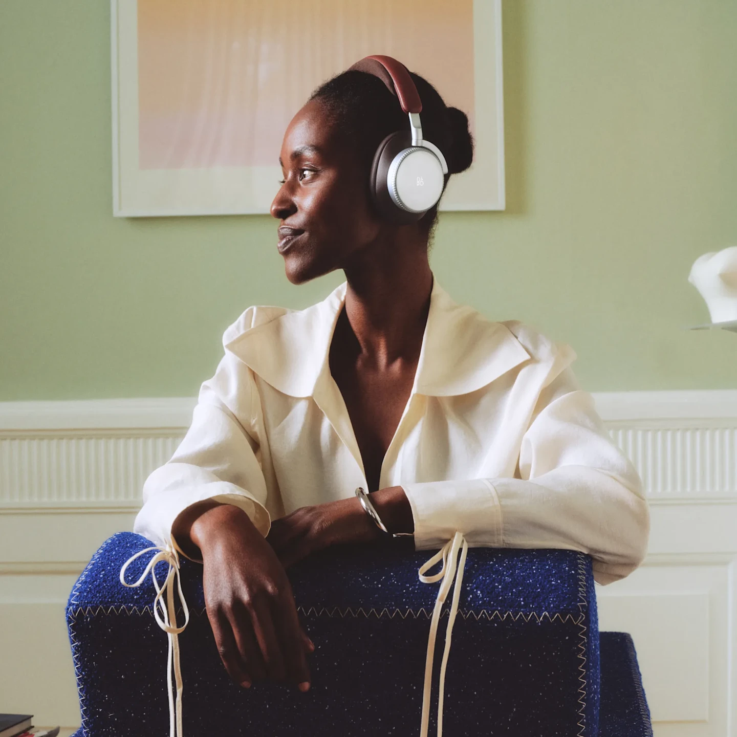 Person wearing brown and silver Beoplay H100 Centennial Colleciton headphones in white shirt leaning on blue cushioned furniture against light green wall.