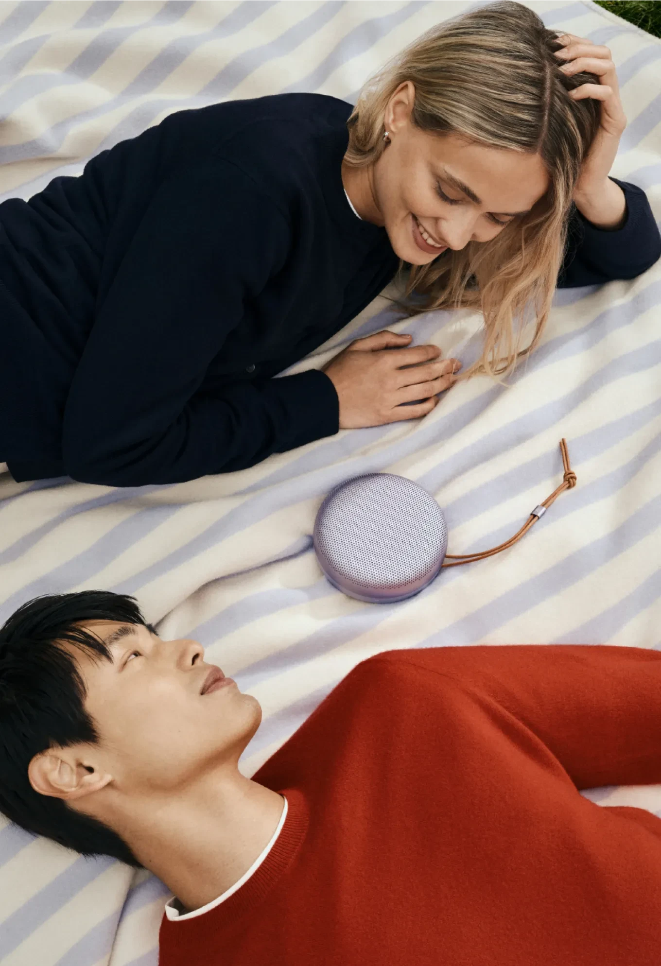 A couple lying face-to-face on a picnic mat with a Bang & Olufsen Beosound A1 in Lavender placed between them, sharing a relaxed moment through music.