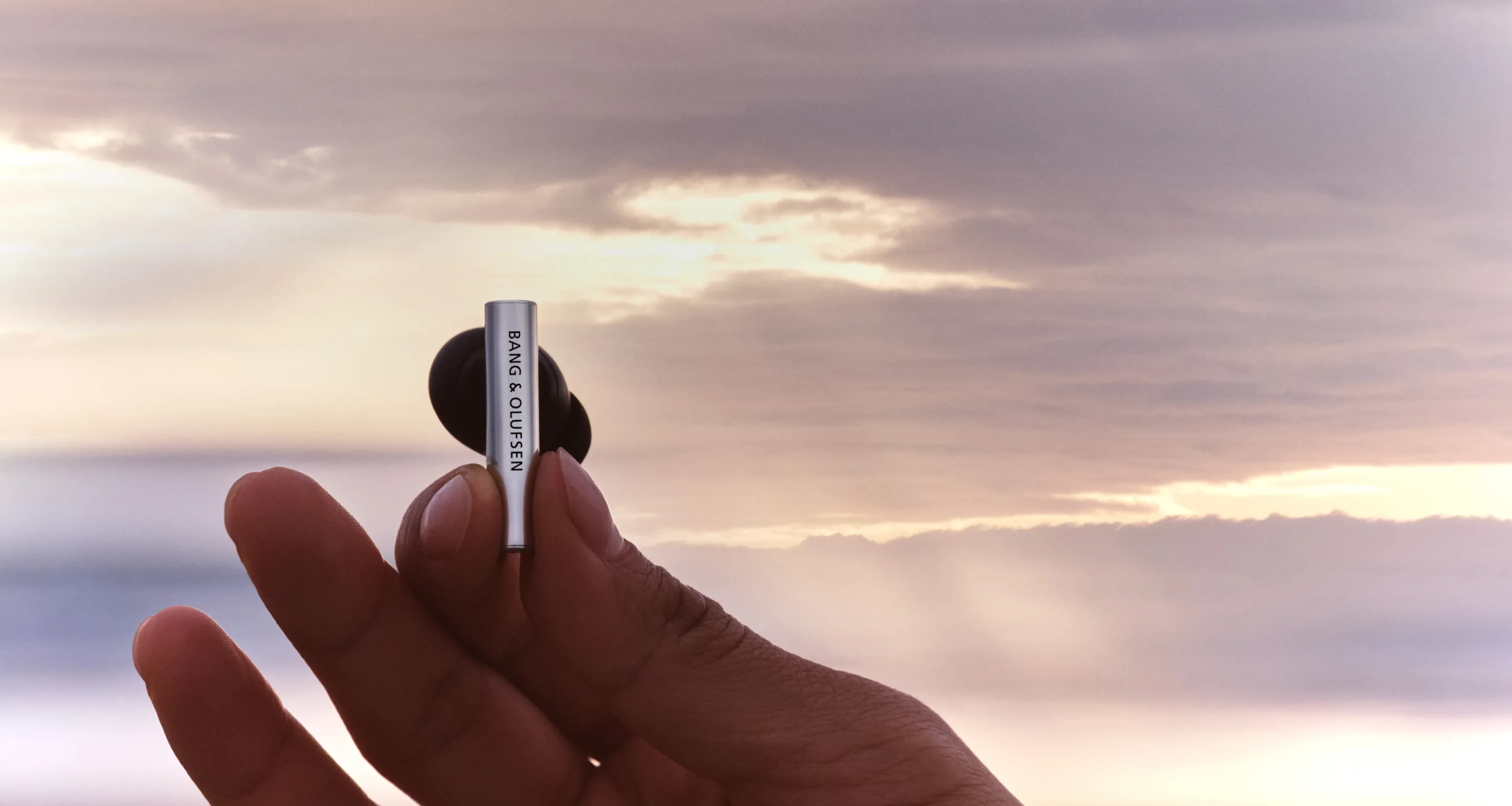 A hand holding one of Beo Grace earphone piece with a beautiful sunset background behind