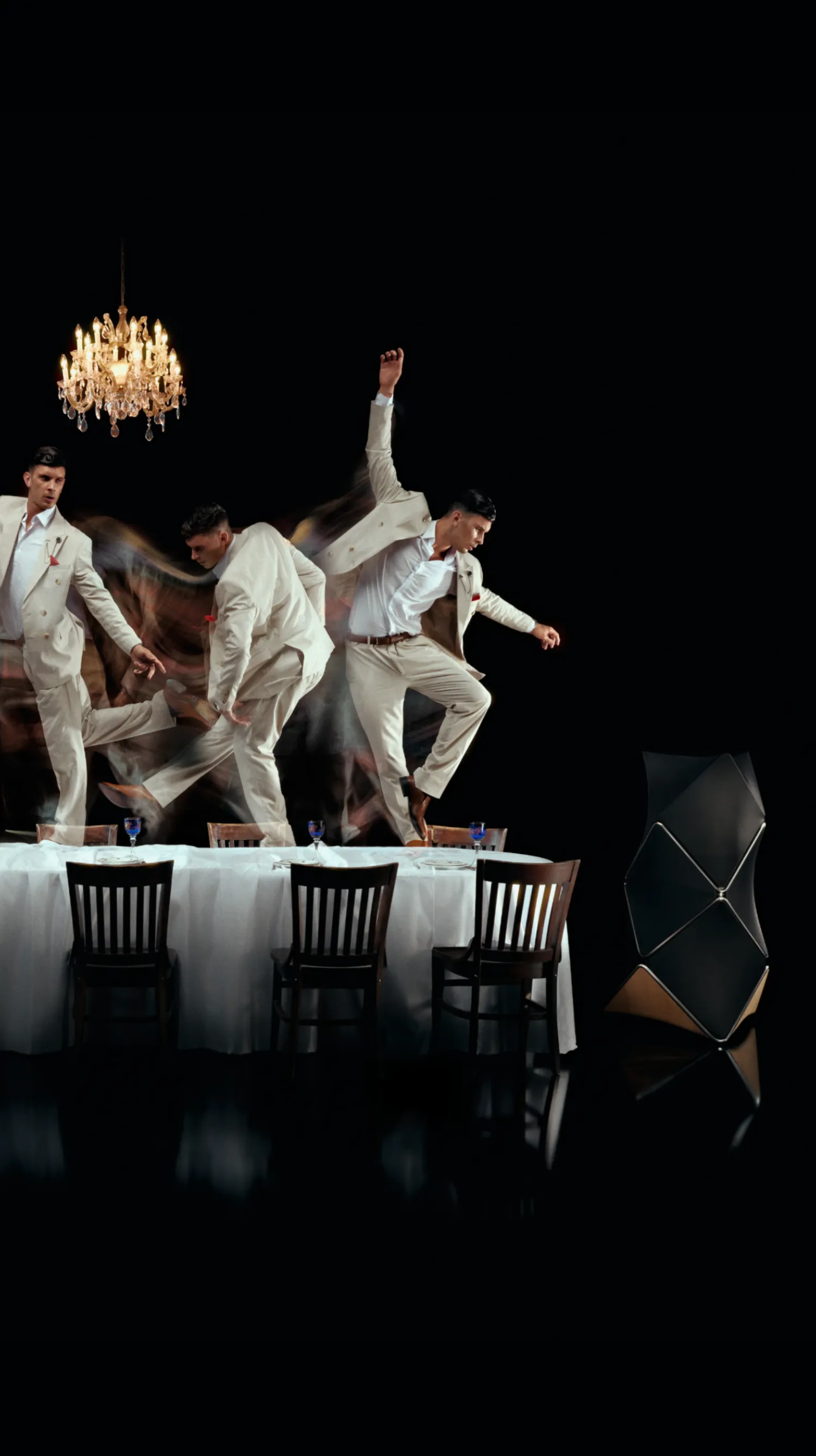 Dancers in white suits performing on a dining table under a crystal chandelier, creating motion blur against a dark background.