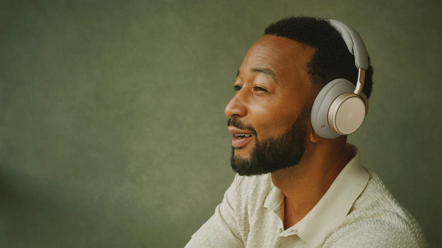 John Legend depicted wearing Bang & Olufsen Beoplay H100 over-ear headphones in the Hourglass Sand colorway, photographed against a green background in a relaxed, music-listening pose.